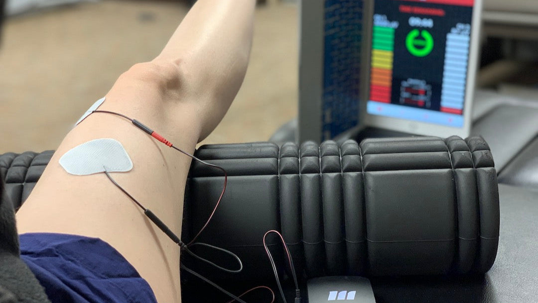 Person using a TENS unit on their arm with a computer monitor displaying data in the background.
